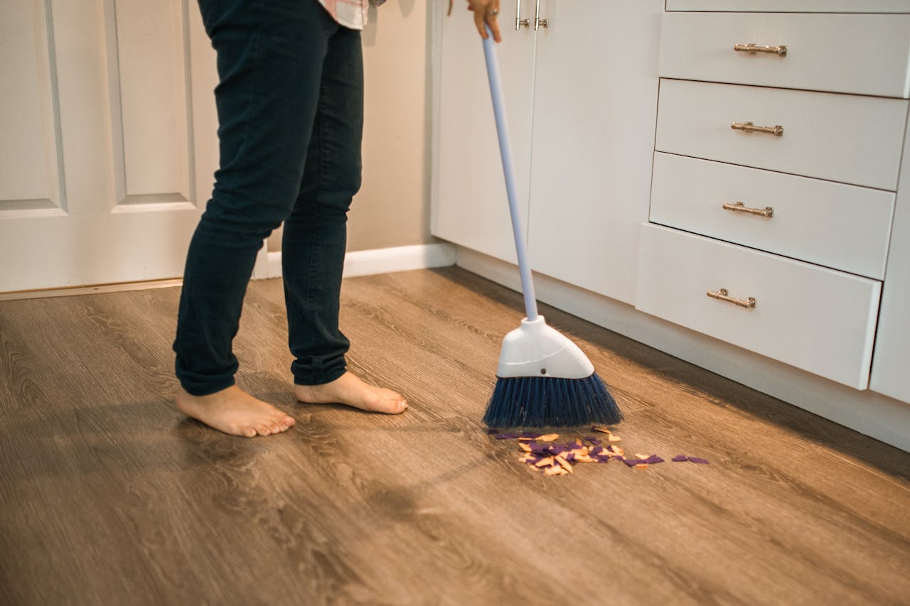 Person sweeping wooden kitchen floor with a broom, showing daily cleaning routine.